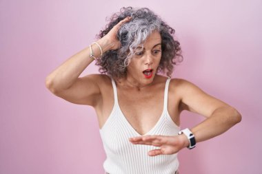 Middle age woman with grey hair standing over pink background looking at the watch time worried, afraid of getting late 