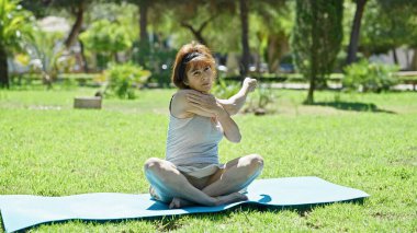 Orta yaşlı bir kadın parkta yoga minderinde oturuyor.