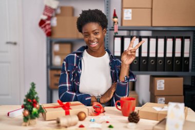 Küçük işletmelerde çalışan Afrikalı Amerikalı bir kadın Noel süslemeleri yapıyor. Kameraya gülümseyen bir gülümsemeyle zafer işareti yapıyor. İki numara.. 