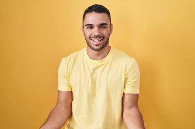 Young hispanic man standing over yellow background smiling cheerful with open arms as friendly welcome, positive and confident greetings 