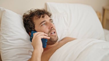 Genç İspanyol adam yatakta üstsüz yatıyor. Akıllı telefondan konuşuyor. Yatak odasına üzgün bakıyor.