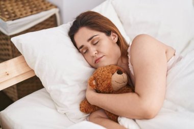 Genç bir kadın oyuncak ayıyı kucaklıyor, yatakta uzanıyor, yatak odasında uyuyor.