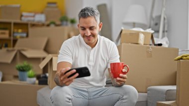 Genç İspanyol adam kahve içiyor ve yeni evinde akıllı telefondan video izliyor.