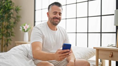 Beyaz saçlı, akıllı telefon kullanan, yatakta oturan bir adam.