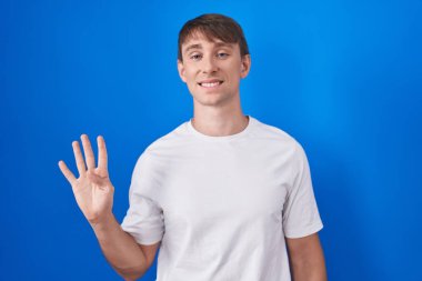 Mavi arka planda duran beyaz tenli sarışın adam dört numaralı parmaklarıyla gülümseyerek kendinden emin ve mutlu görünüyor.. 