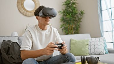 Sanal gerçeklik gözlüğü ve joystick kullanarak evdeki oyun dünyasına dalan, rahat ama konsantre olmuş 3 boyutlu bir yaşam tarzı yaşayan İspanyol asıllı genç.