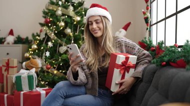 Genç sarışın kadın elinde akıllı telefon hediyesiyle evde Noel ağacının yanında oturuyor.