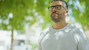 Parkta gözleri kapalı nefes alan gri saçlı bir adam.