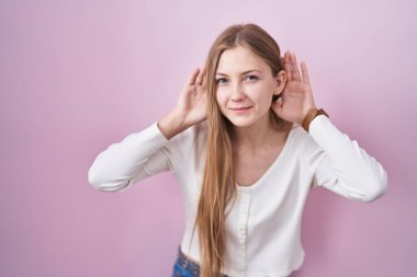 Young caucasian woman standing over pink background trying to hear both hands on ear gesture, curious for gossip. hearing problem, deaf 