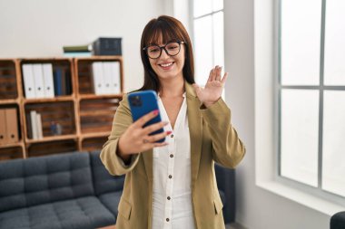 Ofiste akıllı telefonuyla çalışan genç esmer kadın pozitif ve mutlu görünüyor. Dişlerini gösteren kendine güvenen bir gülümsemeyle ayakta duruyor. 