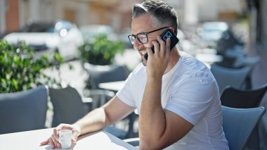 Gri saçlı, kahve içen, kahve dükkanının terasında akıllı telefondan konuşan adam.