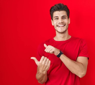 Young hispanic man standing over red background pointing to the back behind with hand and thumbs up, smiling confident 