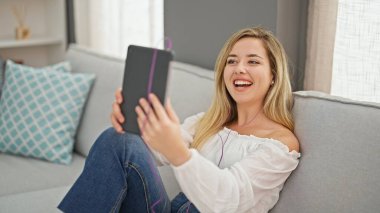 Young blonde woman having video call sitting on sofa at home