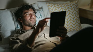 Young hispanic man using touchpad lying on bed at bedroom