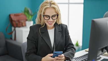 Ofiste çalışan sarışın iş kadını akıllı telefon kullanıyor.