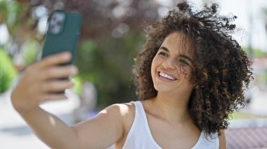 Genç, güzel, İspanyol bir kadın gülümsüyor. Parktaki akıllı telefonun yanında özçekim yapıyor.