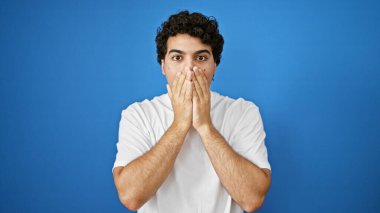İzole edilmiş mavi arka planda ağzını kapatan sürpriz bir ifadeyle genç latin adam.