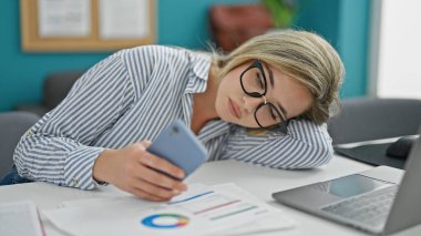 Genç sarışın iş kadını ofiste akıllı telefon kullanarak sıkılmış ve yorgun çalışıyor.