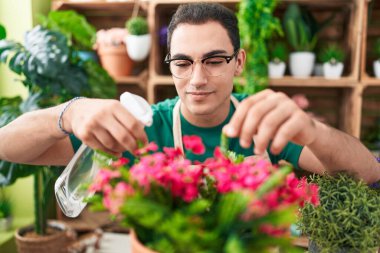 Young hispanic man florist using diffuser watering plant at flower shop