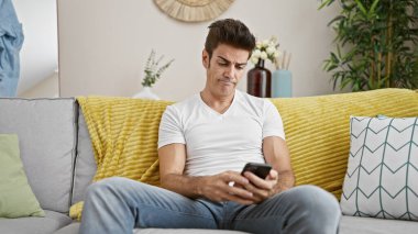 Genç İspanyol adam evde ciddi bir ifadeyle akıllı telefon kullanıyor.