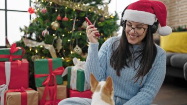 Chihuahua köpeği olan genç İspanyol kadın evde kulaklık ve Noel şapkası takıp müzik dinliyor.
