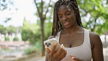 Parkta gülümseyen akıllı telefon kullanan Afrikalı Amerikalı kadın.