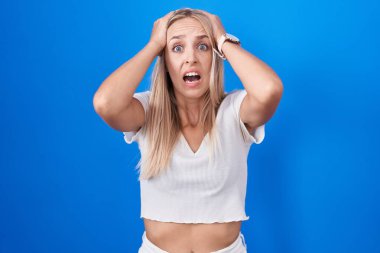 Young caucasian woman standing over blue background crazy and scared with hands on head, afraid and surprised of shock with open mouth 