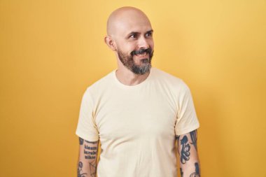 Sarı arka planda dikilen İspanyol dövmeli adam yüzünde gülümseme, doğal bir ifade ile yan yana bakıyor. Kendinden emin gülen. 