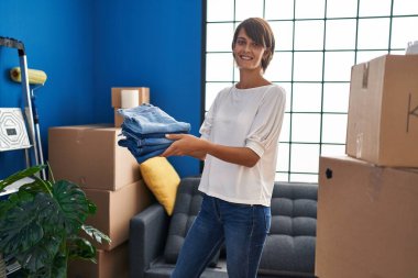 Young beautiful hispanic woman smiling confident holding folded jeans at new home