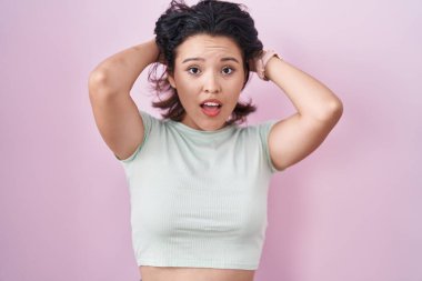 Pembe arka planda duran İspanyol genç kadın deli ve elleri başının üstünde korkmuş, korkmuş ve ağzı açık şoktan şaşırmış. 