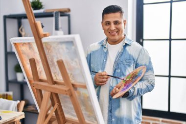 Sanat stüdyosunda kendine güvenen genç Latin sanatçı gülümsüyor.