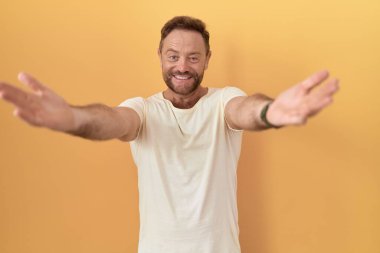 Sarı arka planda sakallı orta yaşlı bir adam kameraya sarılmak için gülümseyerek bakıyor. Mutluluğu kucaklayan neşeli bir ifade.. 