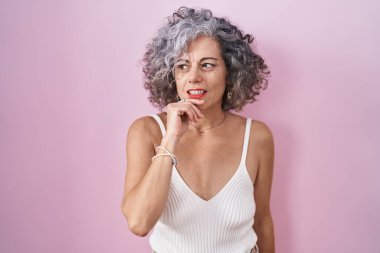 Middle age woman with grey hair standing over pink background with hand on chin thinking about question, pensive expression. smiling and thoughtful face. doubt concept. 