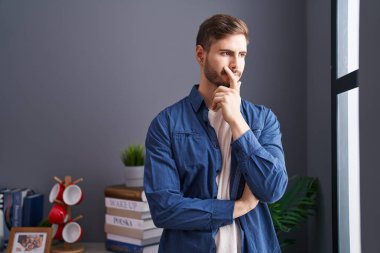 Kolları çaprazlanmış genç, beyaz bir adam evinde duruyor.