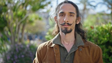 Young hispanic man standing with serious expression at park