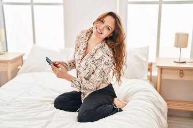 Genç, güzel, İspanyol bir kadın yatakta akıllı telefon kullanıyor.