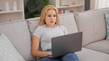 Evdeki kanepede dizüstü bilgisayar kullanan genç sarışın kadın.
