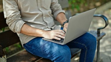 Genç İspanyol adam parkta bankta dizüstü bilgisayar kullanıyor.