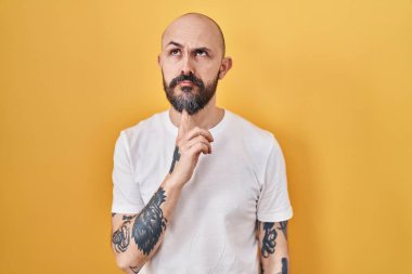 Sarı arka planda duran İspanyol dövmeli genç adam şüpheye odaklanmıştı. Parmakları çenesinde ve yukarı bakıyordu. 