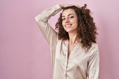 Hispanic woman with curly hair standing over pink background smiling confident touching hair with hand up gesture, posing attractive and fashionable 