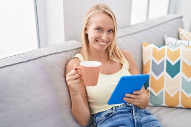 Genç sarışın kadın kahve içiyor ve evde dokunmatik ped kullanıyor.