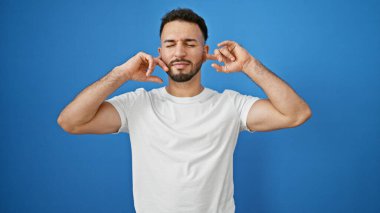 Genç Arap adam, kulaklarıyla kulaklarını tıkayarak izole edilmiş mavi arka planda gürültü yapıyor.