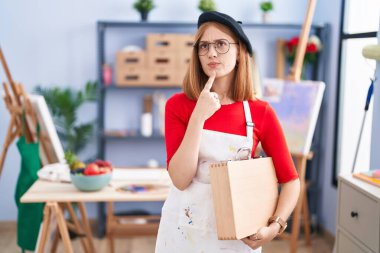Sanat stüdyosundaki genç kızıl saçlı kadın elinde bir sanat çantası tutuyordu. Düşünceleri şüpheye odaklanmıştı. Parmakları çenesinde ve yukarı bakıyordu. 