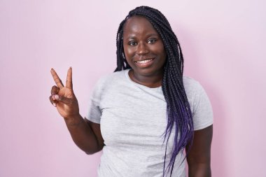 Pembe arka planda duran Afrikalı genç bir kadın iki numarayı gösterip işaret ederken kendinden emin ve mutlu bir şekilde gülümsüyor.. 