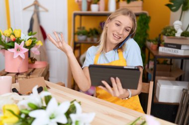 Young blonde woman florist talking on smartphone using touchpad at flower shop