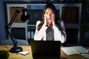 Genç esmer kadın geceleri ofiste dizüstü bilgisayarla çalışıyor. Korkmuş ve şok olmuş, şaşırmış ve şaşırmış bir halde. 