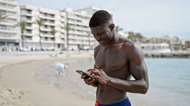 Afrika kökenli Amerikalı turist plajda üstsüz bir şekilde akıllı telefon kullanıyor.
