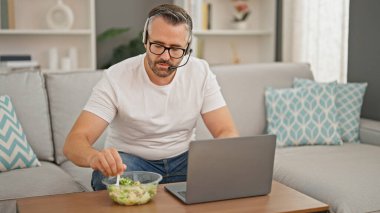 Evde dizüstü bilgisayar ve kulaklık kullanarak salata yiyen gri saçlı adam.