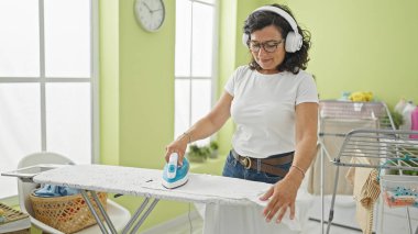 Orta yaşlı İspanyol kadın çamaşırhanede müzik dinliyor. Kıyafetleri ütülüyor.