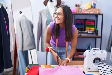 Young beautiful hispanic woman tailor smiling confident cutting cloth at tailor shop